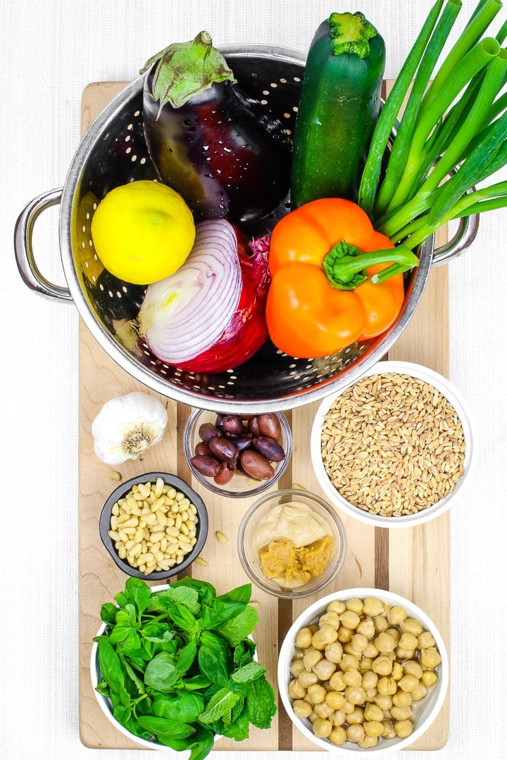 Ingredients on a cutting board. Roast vegetable salad with eggplant, orange peppers zucchini, olives, red onions, garlic, green onions, farro, pine nuts, chickpeas, basil, and lemon.