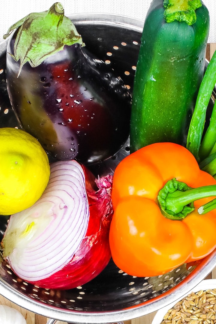 close up of eggplant, lemon, red onion half, orange bell pepper and zucchini in a stainless steel colander.