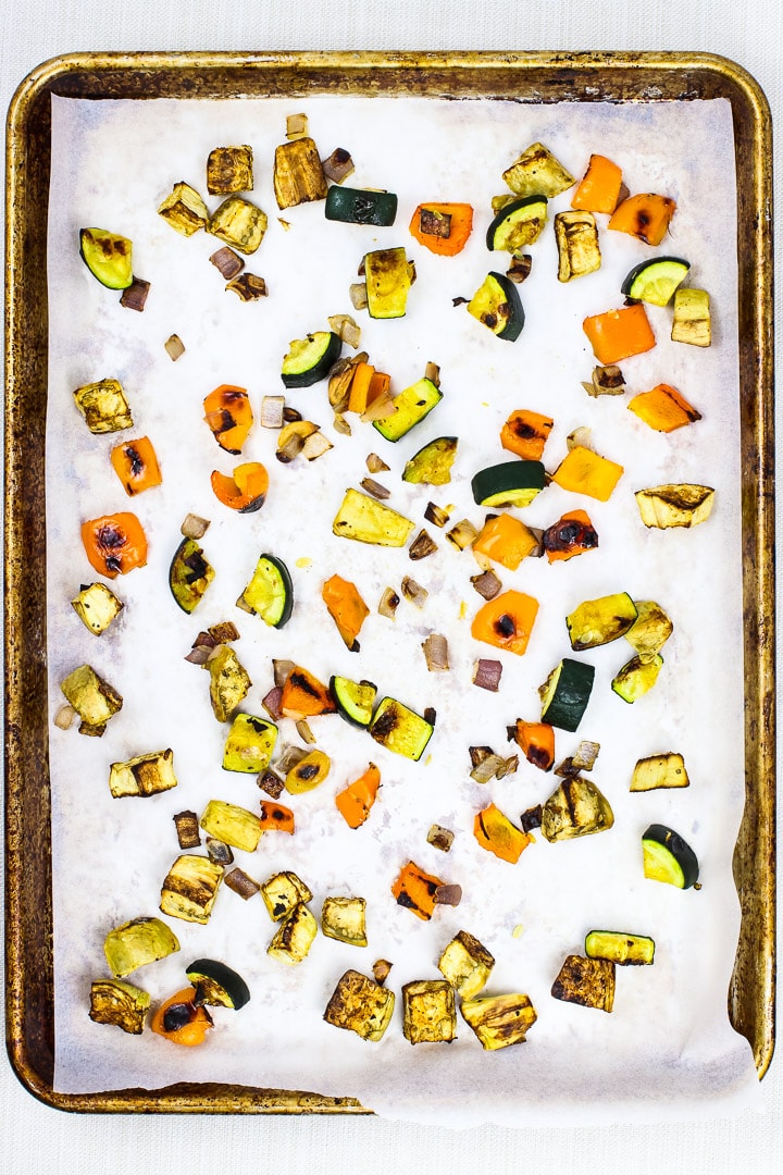 Parchment paper lined baking sheet with pieces of roasted eggplant, zucchini, orange bell pepper, red onion.