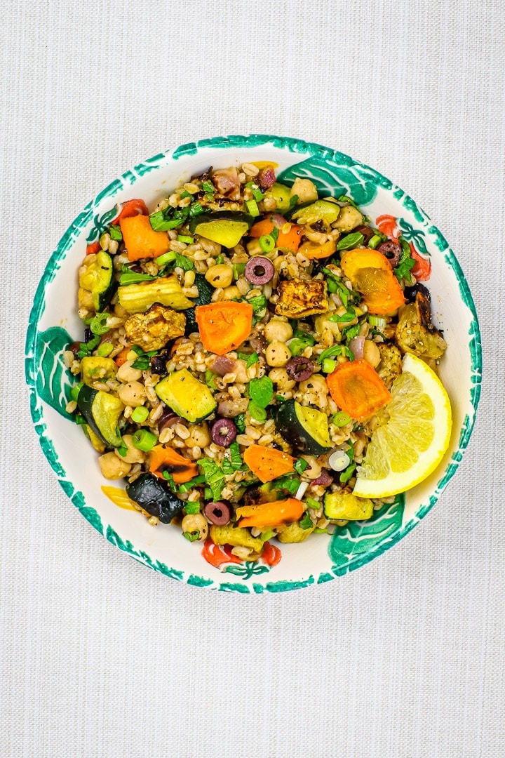 Roast vegetable salad with eggplant, orange peppers zucchini, olives, red onions, sliced green onions, farro, pine nuts, and lemon slice in a leaf patterned bowl. 