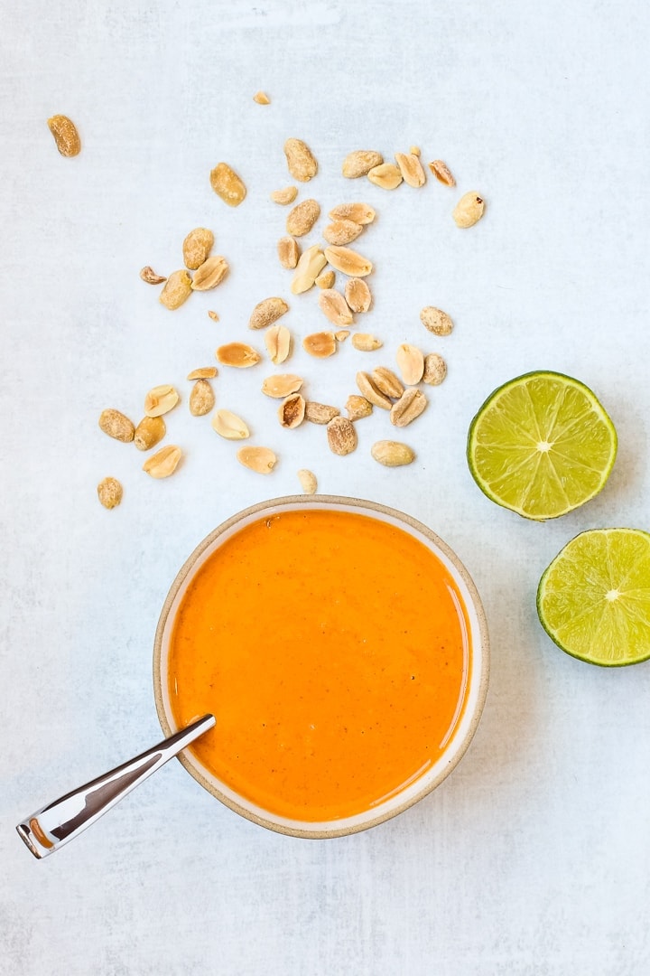 Bowl of thai curry peanut sauce with spoon, cut limes and peanuts on a gray background. 