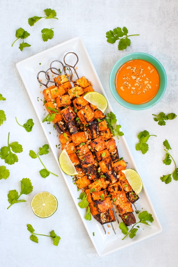 White platter with skewers of grilled eggplant, tofu, pineapple and yellow squash with Thai Curry peanut sauce in a teal bowl on a gray background. Garnished with cilantro, crushed peanuts and lime wedges.