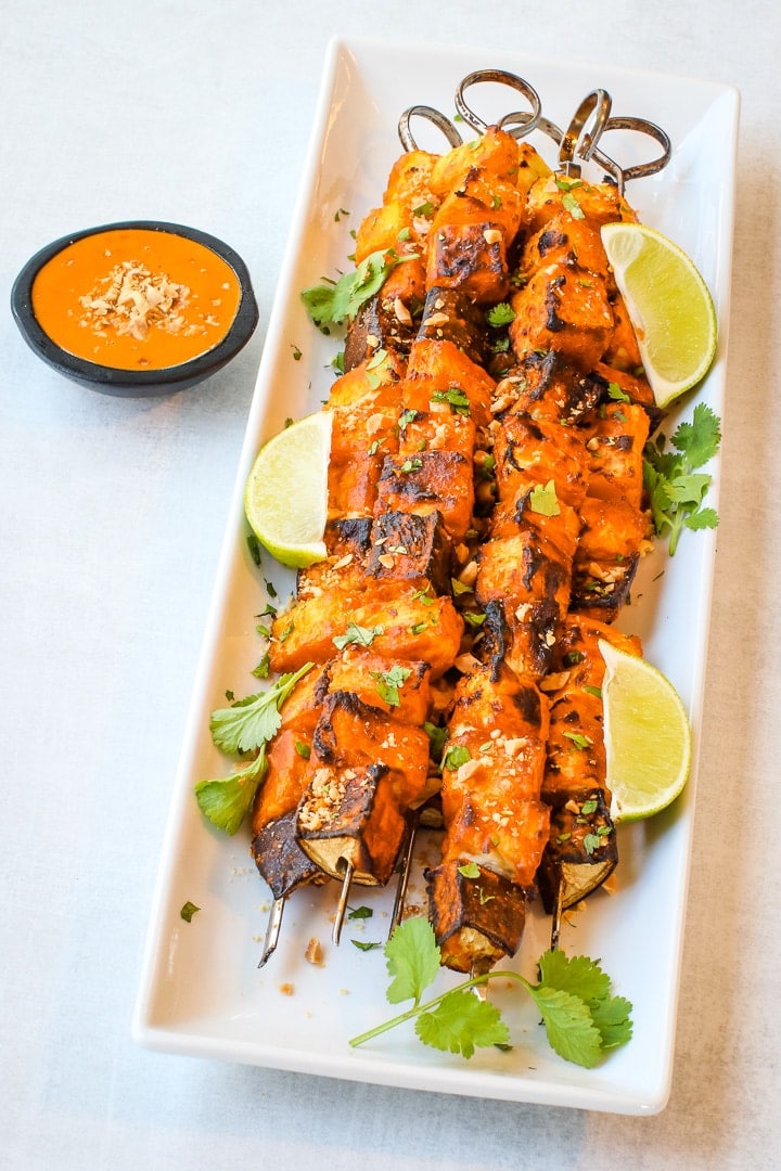 White platter with vegan kebab skewers of grilled eggplant, tofu, pineapple and yellow squash with Thai Curry peanut sauce in a black bowl on a gray background. Garnished with cilantro, crushed peanuts and lime wedges.