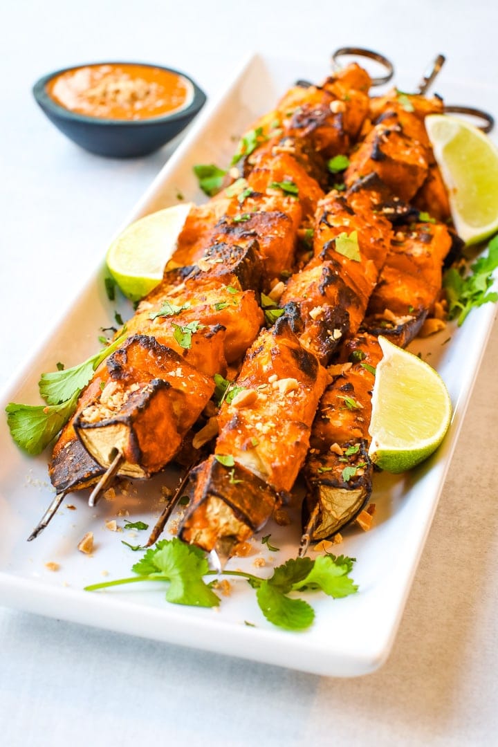 Close up of white platter with vegan kebabs of grilled eggplant, tofu, pineapple and yellow squash with Thai Curry peanut sauce in a black bowl on a gray background. Garnished with cilantro, crushed peanuts and lime wedges.