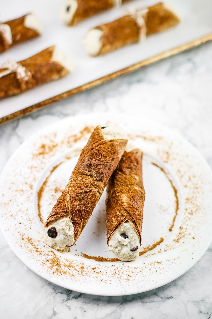 Baked tortilla cannolis coated in coconut sugar and filled with almond ricotta and chocolate chips on a white plate on marble. Tray of connolis in background.