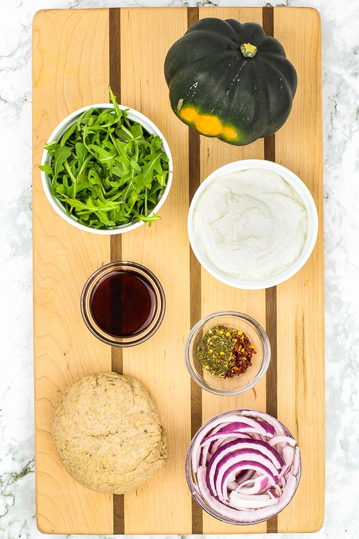 Acorn squash, arugula, almond ricotta, red onion, and pizza dough on a cutting board on marble.