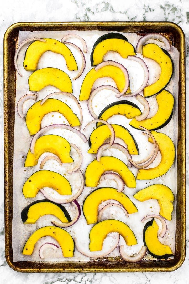 Acorn squash and red onion slices on parchment paper lined baking sheet on marble.