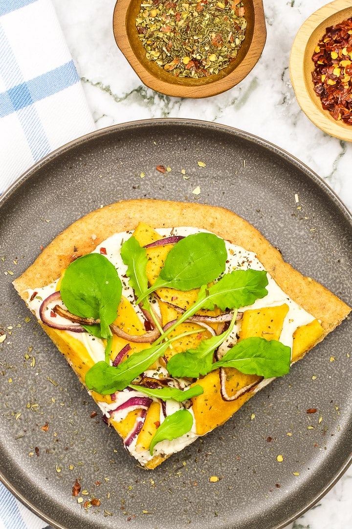 roasted squash white pizza topped with arugula on a gray plate. Little dishes of Italian seasonings and cursed red peppers, blue plaid towel and marble.