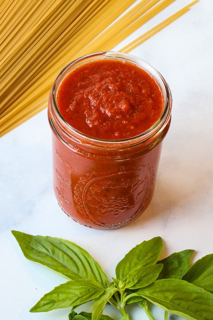 Mason jar filled with tomato sauce, with basil leave and dry spaghetti on white marble. 