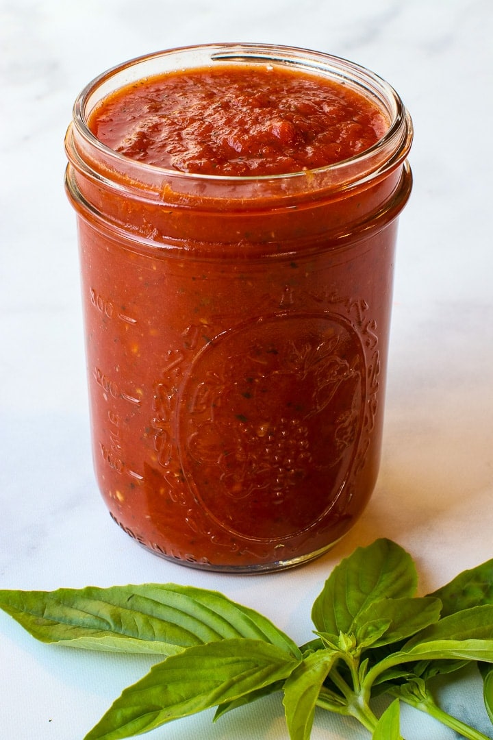 Close up of Mason jar filled with tomato sauce, with basil leave and dry spaghetti on white marble. 