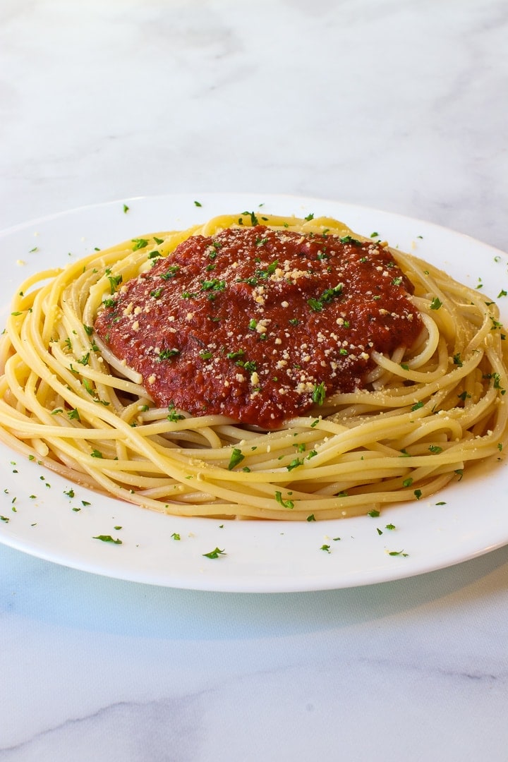 White plate of spaghetti with tomato sauce garnished with vegan parmesan and minced parsley on marble. 