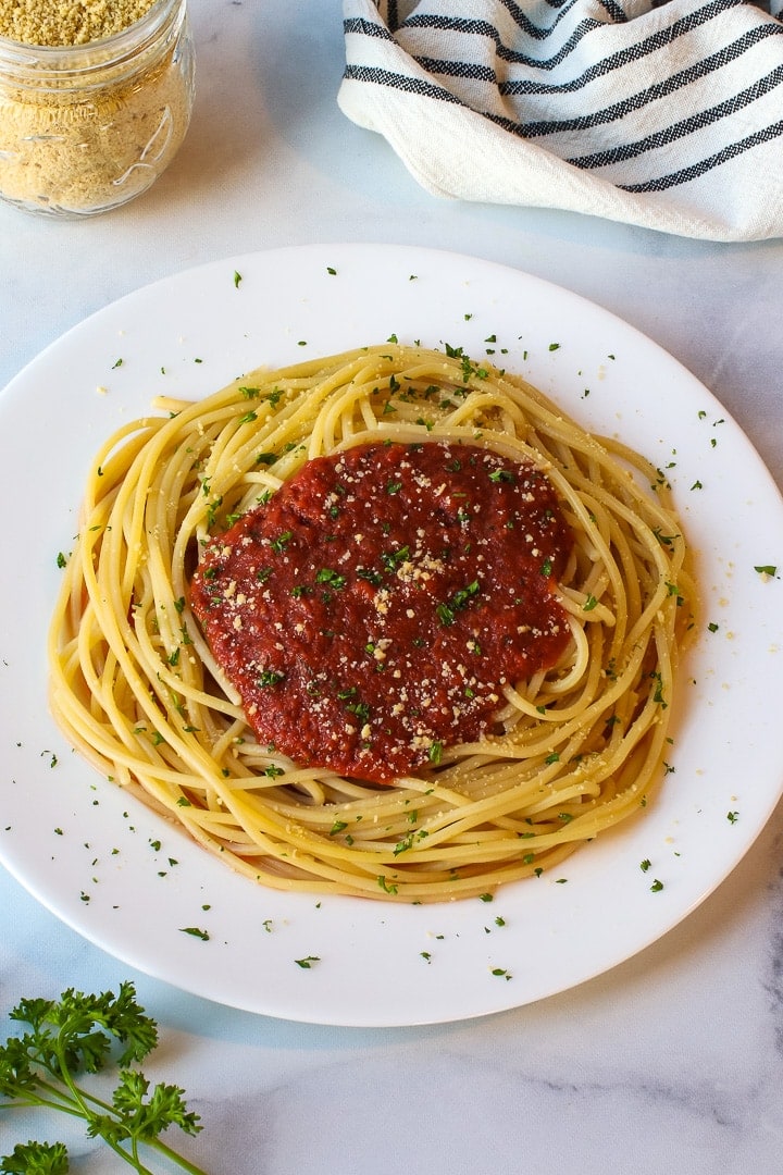 White plate of spaghetti with tomato sauce garnished with vegan parmesan and minced parsley on marble with vegan parm, parsley and a white towel with black stripes. 