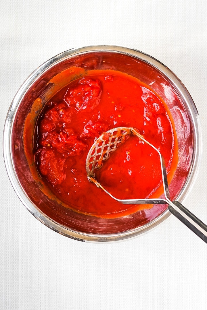 crushing whole canned tomatoes in a stainless steel bowl with a potato masher
