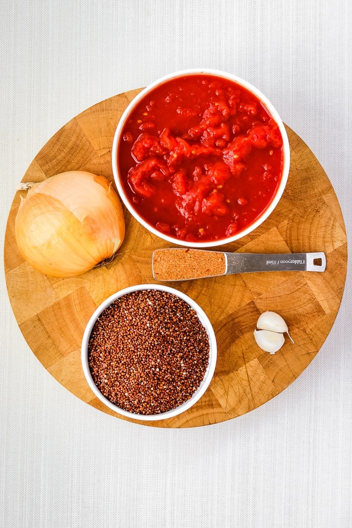 round cutting board with dish of canned tomatoes and red quinoa, with half an onion, 2 garlic cloves and a tablespoon of taco seasoning.