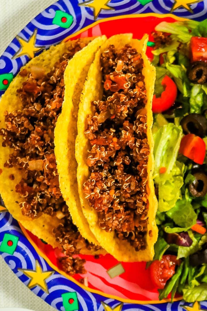 Close up of quinoa taco meat in corn tortilla shells on a festive paper plate with a die of lettuce, diced tomatoes and slices black olives.