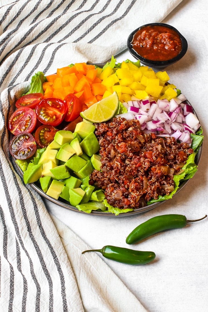 Taco salad with lettuce, halved cherry tomatoes, diced red & yellow bell pepper, diced red onion, avocados cubes and quinoa taco meat on a black striped towel with jalapeno pepper and a black dish of red salsa.