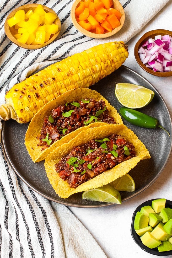 Tacos with red quinoa taco meat in corn tortilla shells on a fray plate with roasted ear of corn, lime and jalapeno, with small dishes of diced yellow and orange peppers, red onion and avocado on a napkin with black stripes. 