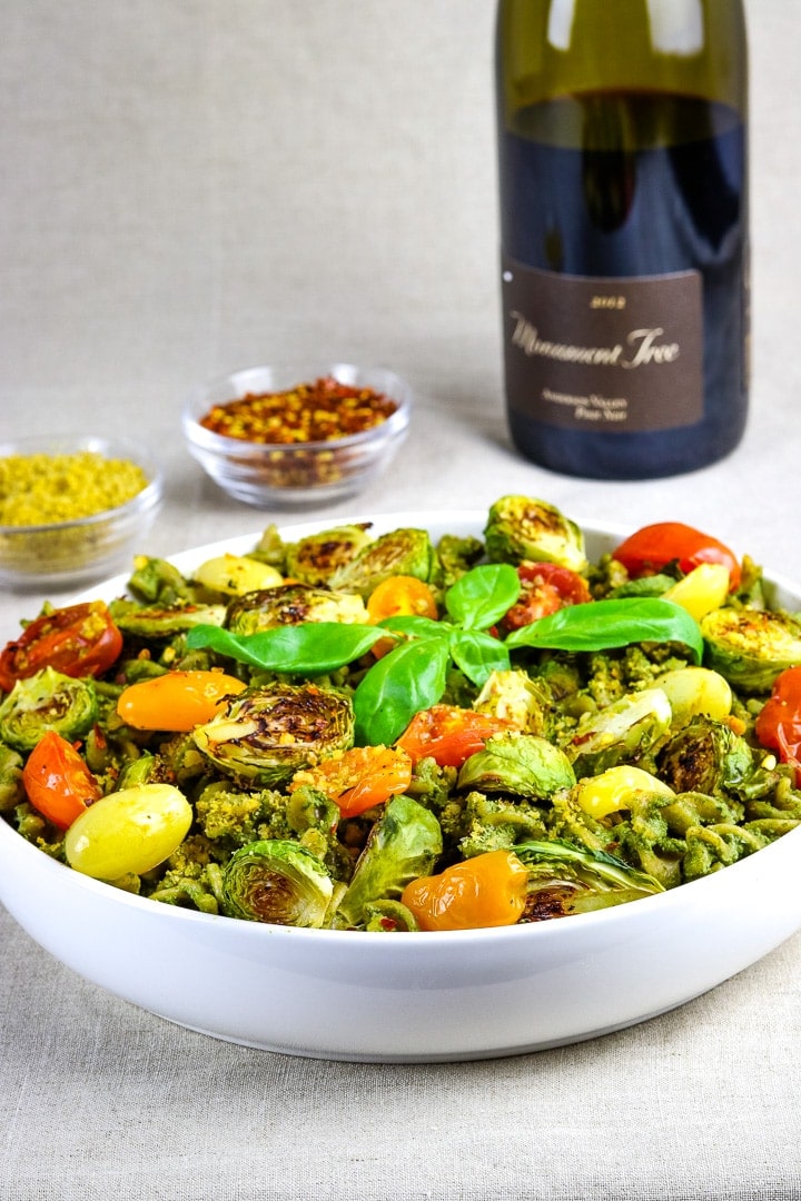 Flat white bowl of curly pasta with pesto sauce, roasted tomatoes and brussles sprouts, topped with basil sprig. Wine bottle an dishes of vegan parmesan and crushed red pepper flakes in background. 