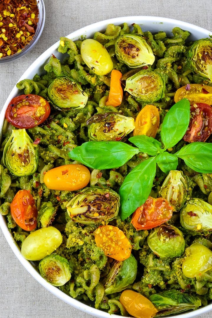 Close up of Flat white bowl of curly pasta with pesto sauce, roasted tomatoes and brussles sprouts, topped with basil sprig. 