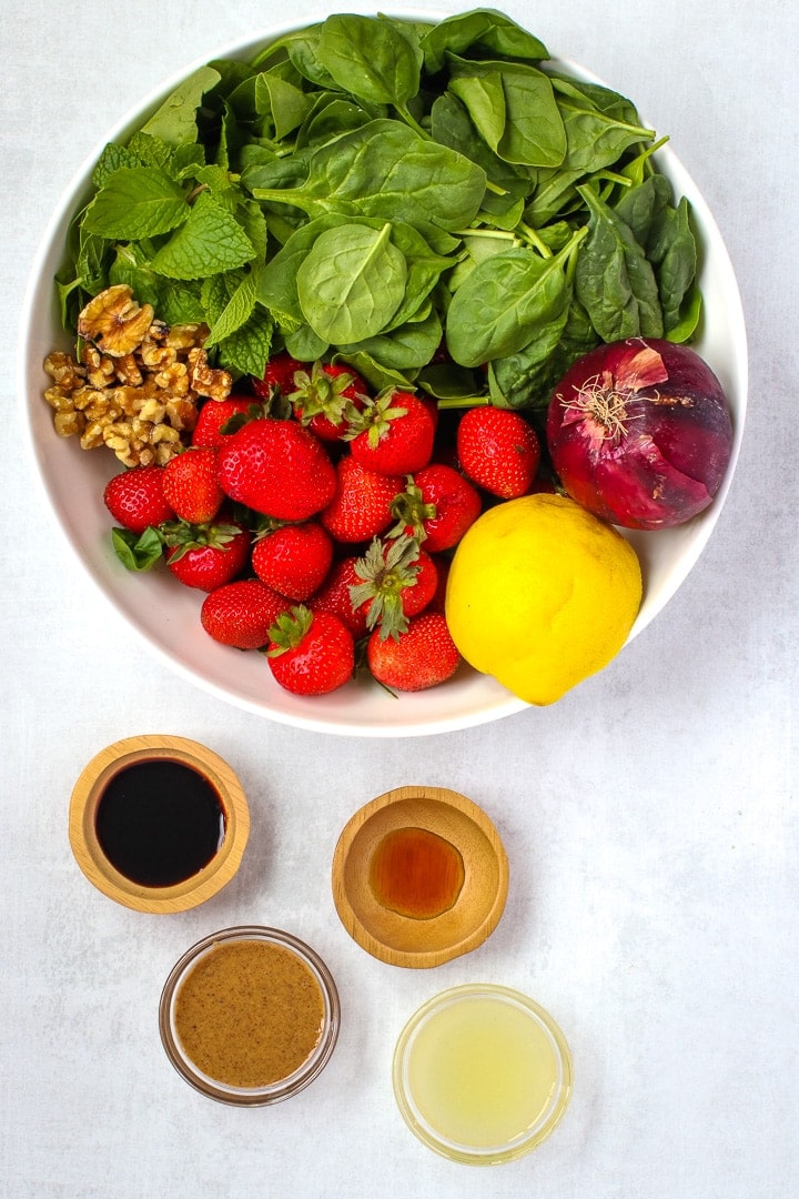Ingredients in a white bowl: spinach, strawberries, whole red onion, whole lemon, walnuts, mint leaves. Small bowls of balsamic vinegar, almond butter, lemon juice, and maple syrup. 