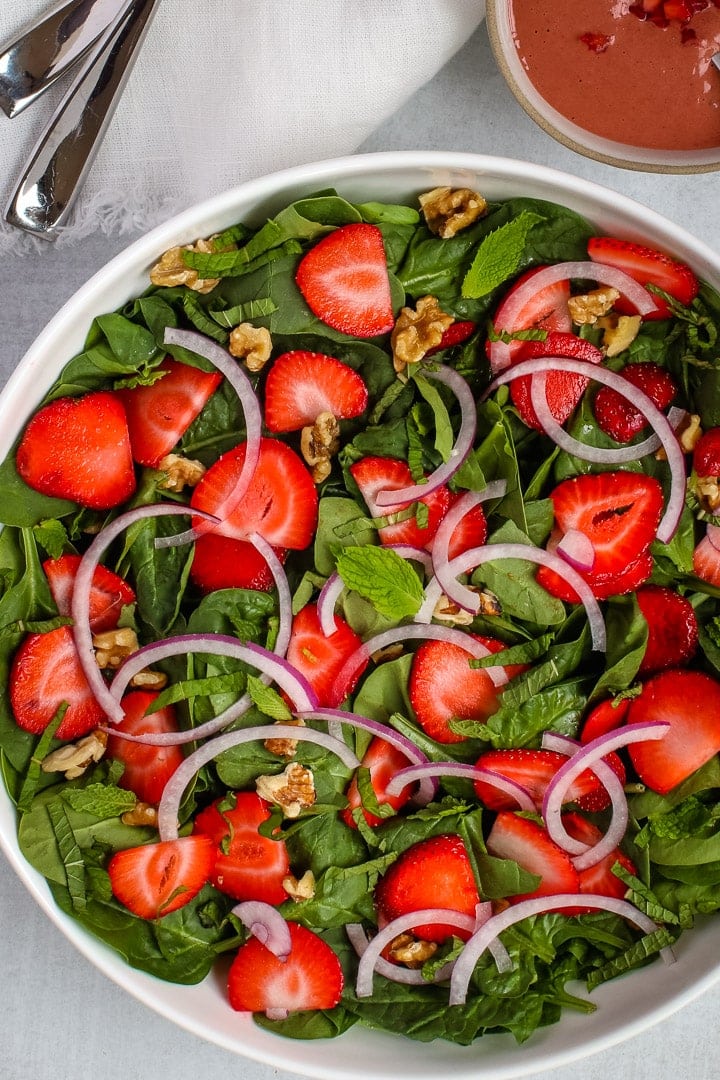 Large white bowl of spinach salad with sliced strawberries, slivered red onions, walnut pieces, fresh mint ribbons and leaves. On a gray background with silverware, a white napkin and strawberry dressing in a bowl. 