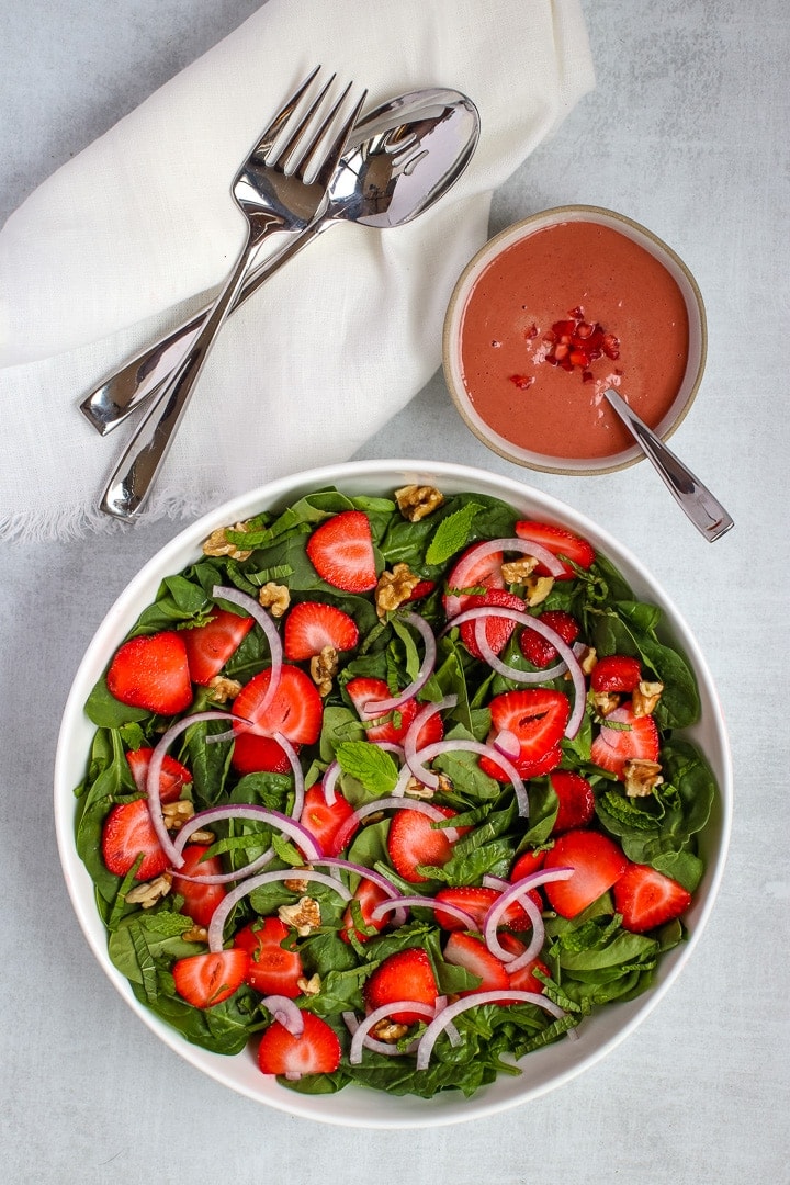 Overhead of Large white bowl of spinach salad with sliced strawberries, slivered red onions, walnut pieces, fresh mint ribbons and leaves. On a gray background with silverware, a white napkin and strawberry dressing in a bowl. 