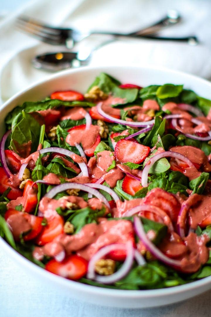 Close up of Large white bowl of spinach salad with sliced strawberries, slivered red onions, walnut pieces, fresh mint ribbons and leaves, with strawberry dressing drizzled on top. On a gray background with silverware on a white napkin. 
