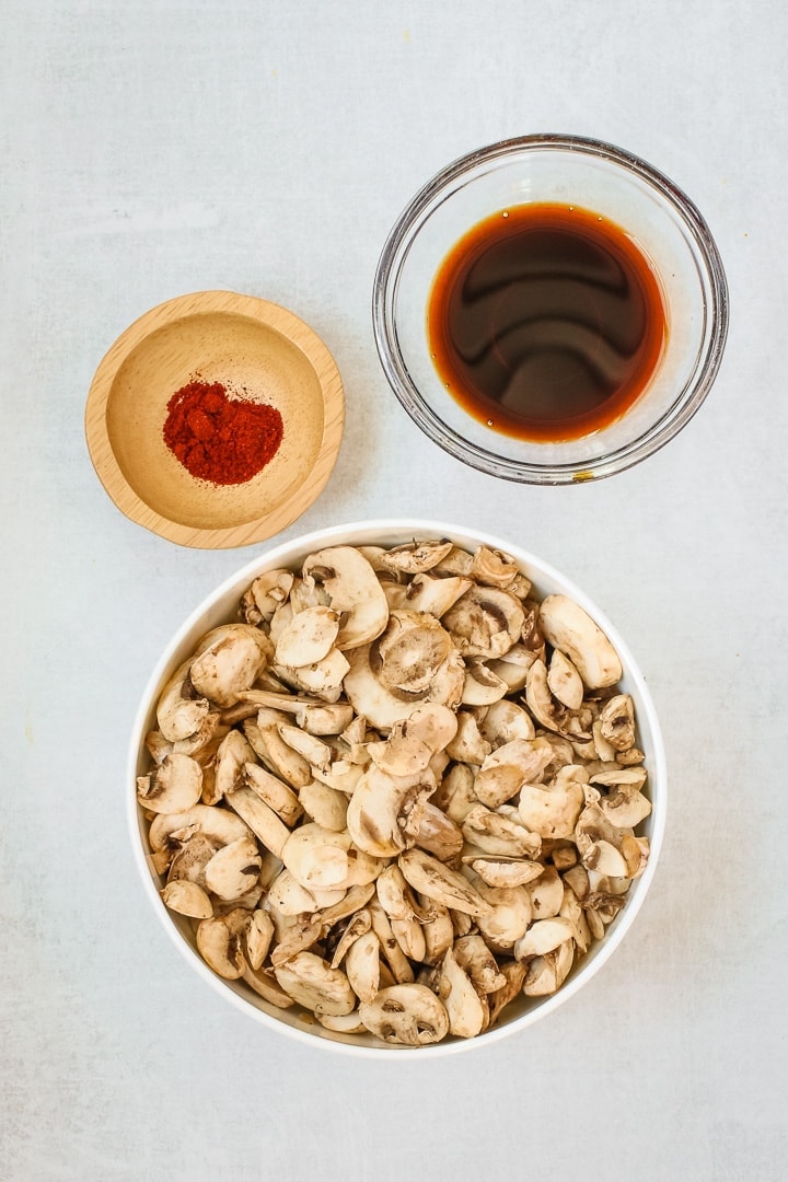 Slices mushrooms, bowl of tamari and liquid smoke, and smoked paprika on a gray background.