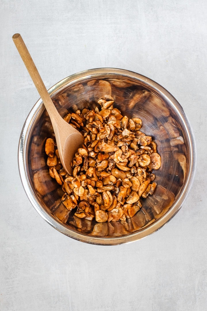 stainless steal bowl of diced mushrooms marinating, with a wooden spoon on a gray background.