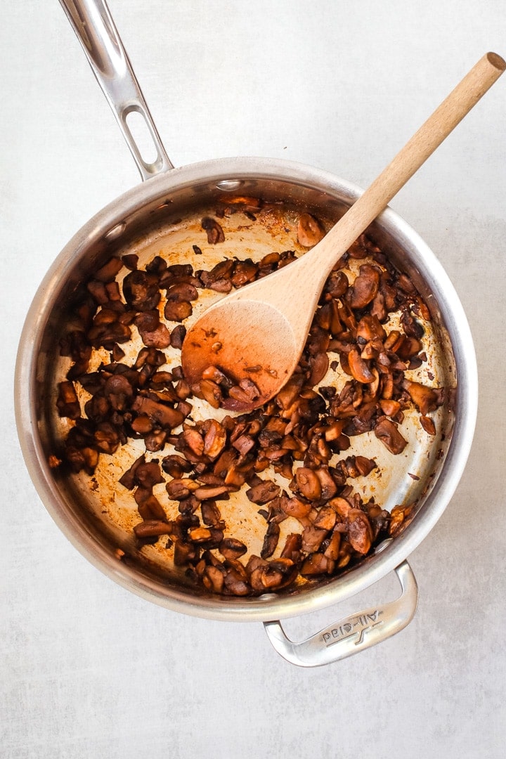 Cooked diced mushrooms in a sauce pan with a wooden spoon on a gray background. 