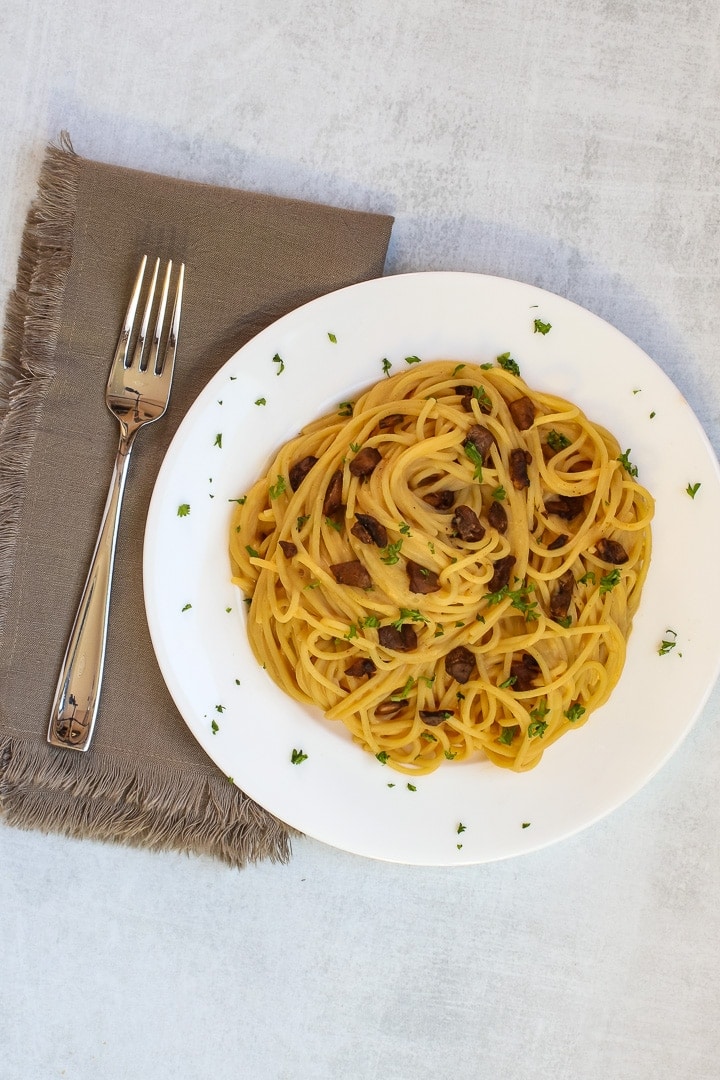 White plate of vegan spaghetti carbonara with mushroom bacon and parsley, with a brown napkin and fork on a gray background.