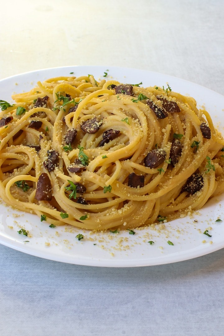 White plate of vegan spaghetti carbonara with mushroom bacon, parsley and vegan parmesan on a gray background.