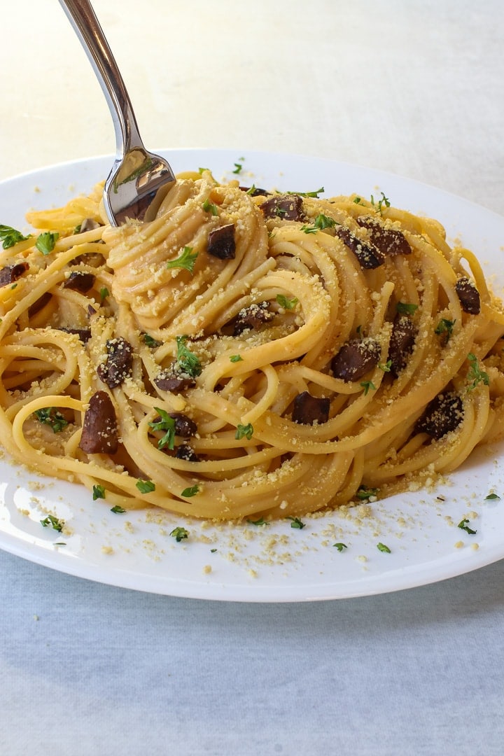 Close up of white plate and fork twirling vegan spaghetti carbonara with mushroom bacon, parsley and vegan parmesan on a gray background.