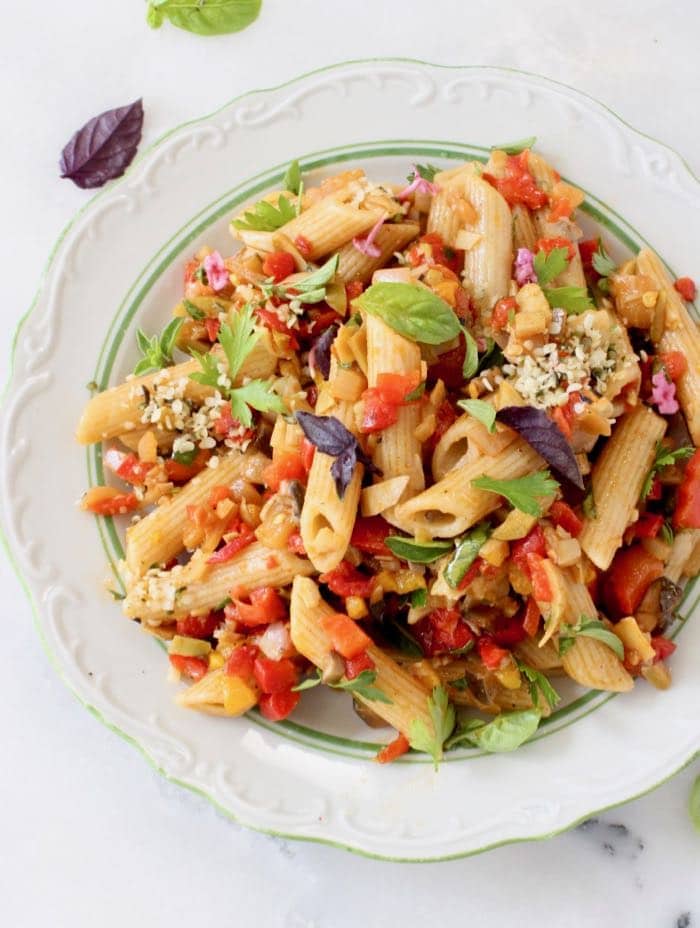 White plate with penne pasta, tomato chunks, green and purple basil leaves.