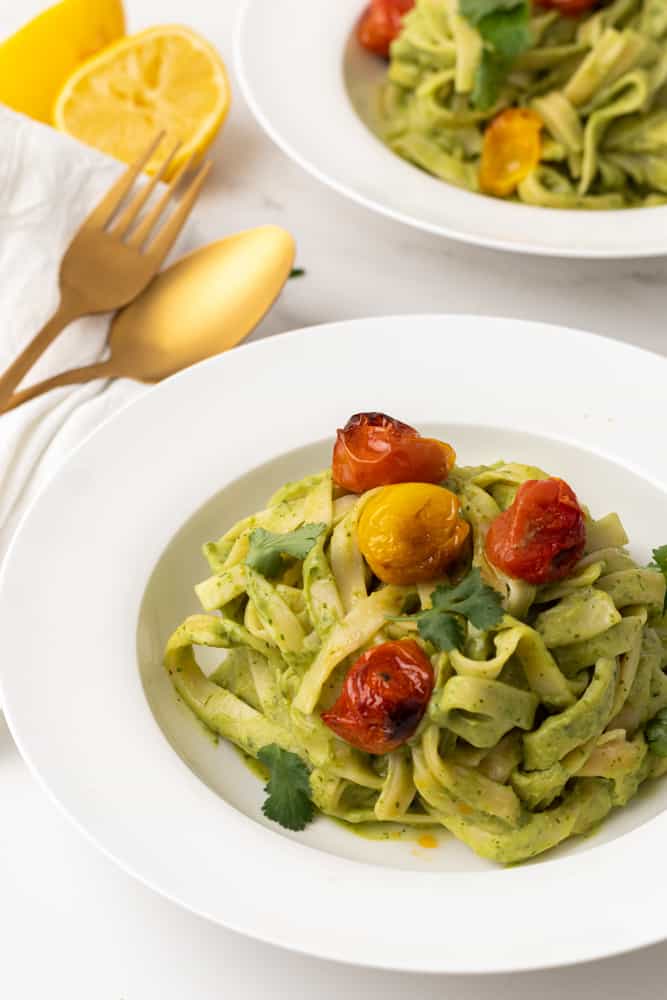 Fettuccine with green pasta sauce and blistered cherry tomatoes in a white dish. Gold knife and fork and lemon in background. 