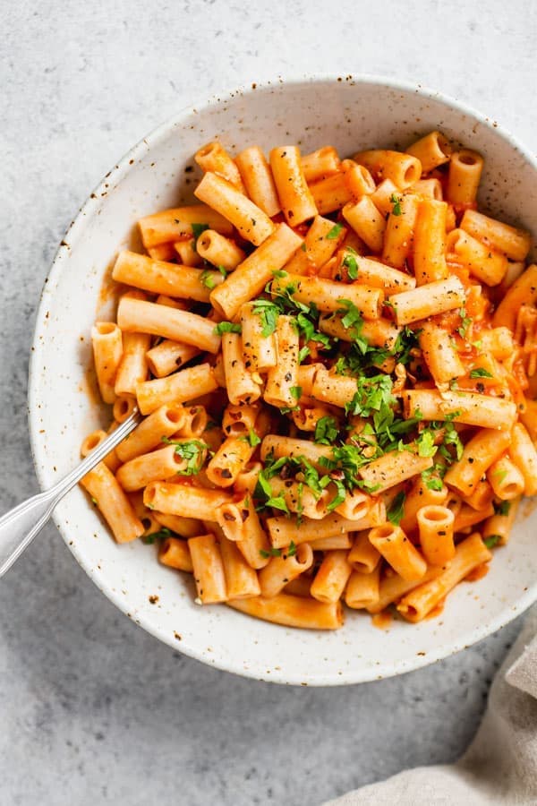 Flecked white bowl with creamy red pepper penne pasta, topped with herbs with fork. 