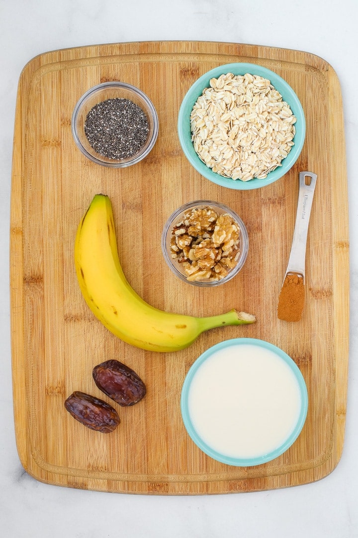 ingredients: rolled oats, plant milk, banana, chia seeds, walnuts, cinnamon and dates on a cutting board.