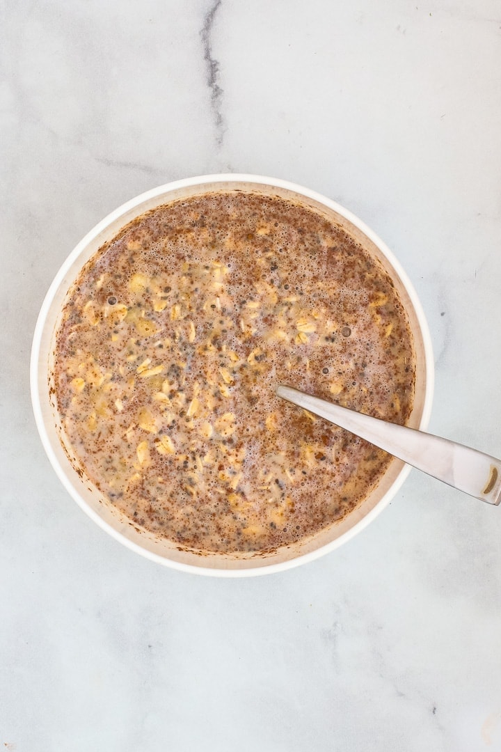 Stirring banana oat mixture together with a spoon in a white bowl on gray marble.