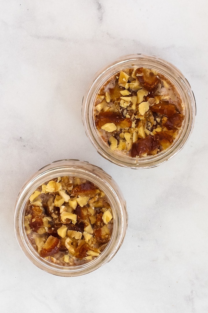 Two mason jars of banana overnight oats topped with chopped walnuts and dates on gray marble.