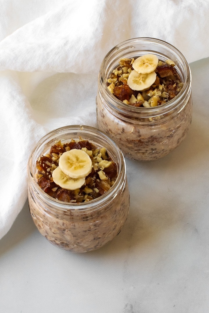 Two small mason jars with banana overnight oats topped with chopped dates and walnuts and banana slices. Pictured with a white towel.