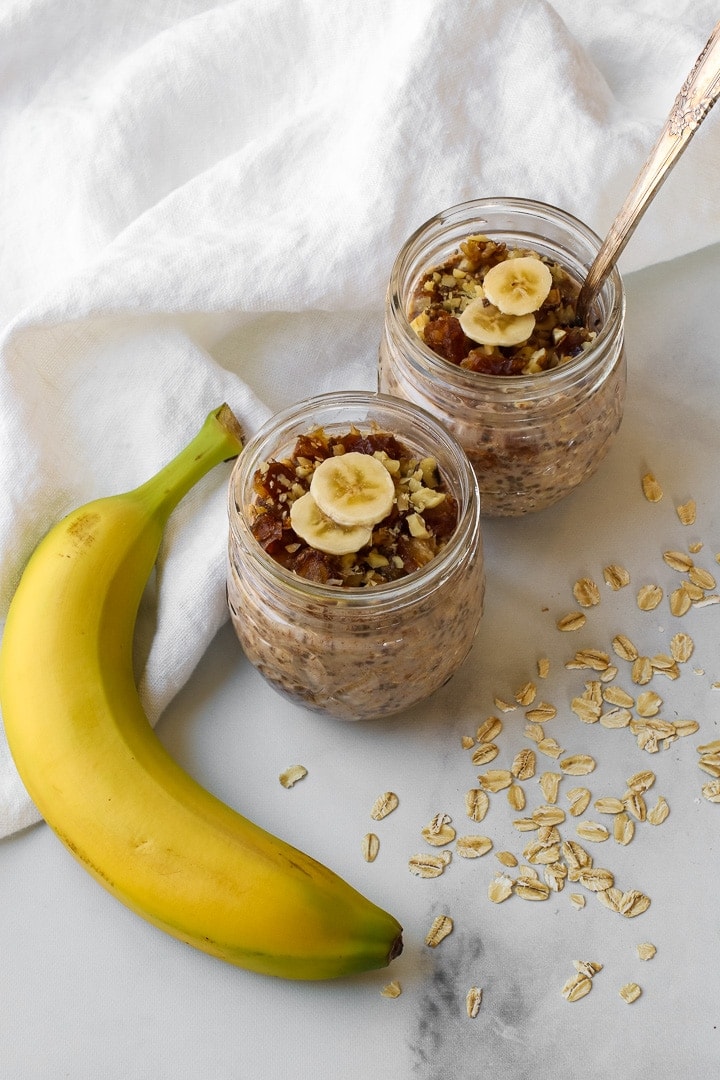 Two small mason jars with banana overnight oats topped with chopped dates and walnuts and banana slices. Pictured With a whole banana, dry oats and white towel.