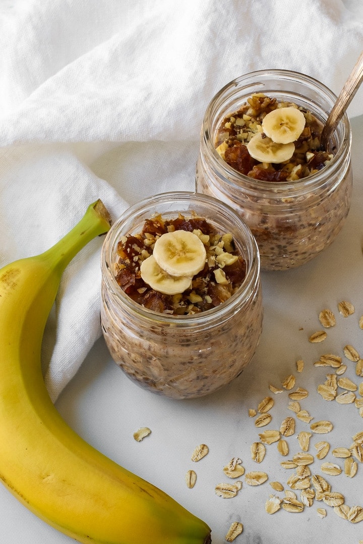 Two small mason jars with banana overnight oats topped with chopped dates and walnuts and banana slices. Pictured With a whole banana, dry oats and white towel.