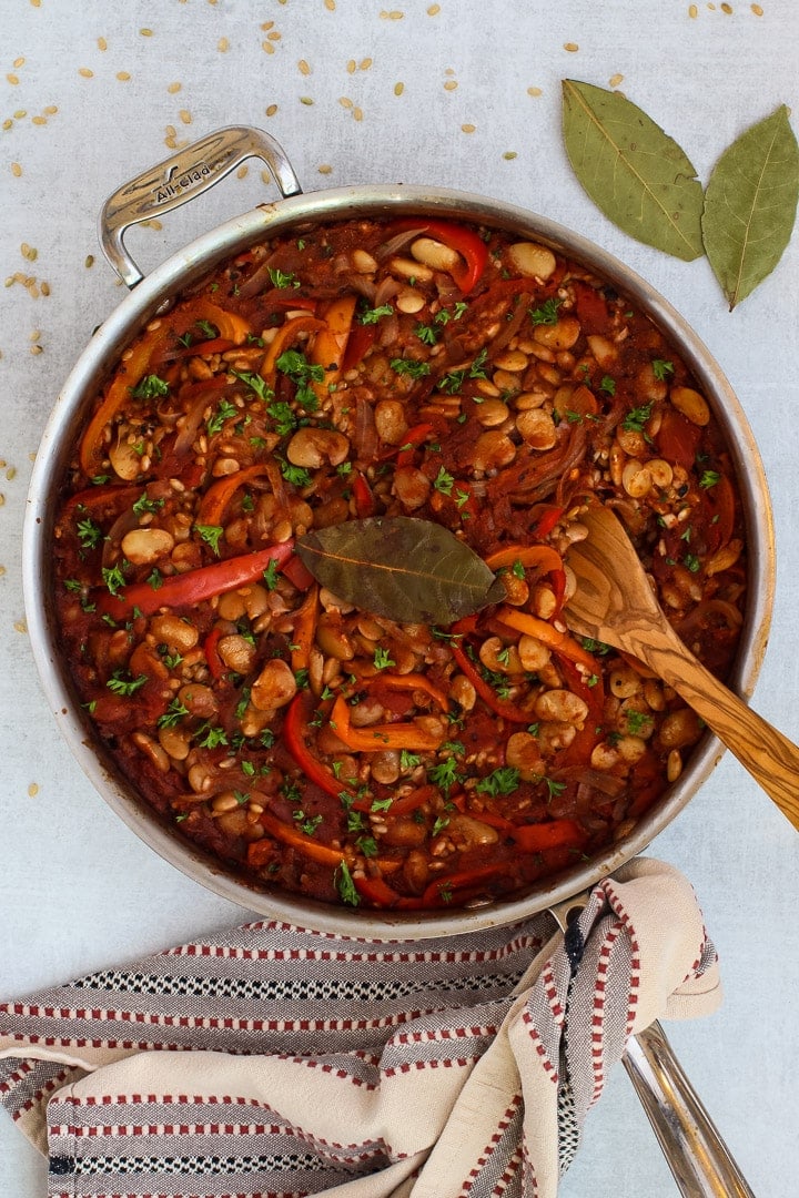 Skillet with Spanish Rice and Beans with slivered onions, red and yellow bell peppers, parsley and bay leaf with red and gray striped towel and wooden spoon.