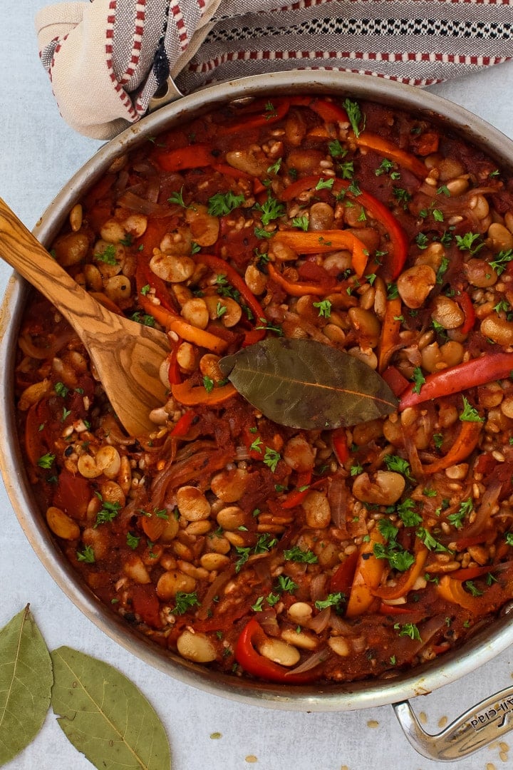 Close up of skillet with Spanish Rice and Beans with slivered onions, red and yellow bell peppers, parsley and bay leaf with red and gray striped towel and wooden spoon.