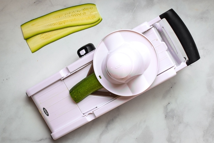 Mandoline slicing a zucchini, with zucchini slices on marble.