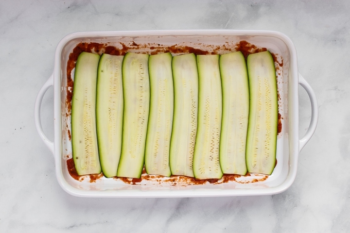 White baking dish with tomato sauce topped with zucchini slices on gray marble. 