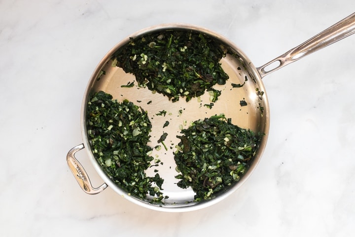 Skillet with cooked spinach, basil, garlic, and onion on marble.