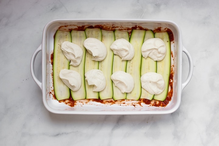 White baking dish with tomato sauce topped with zucchini slices and dollops of almond ricotta cheese on gray marble. 