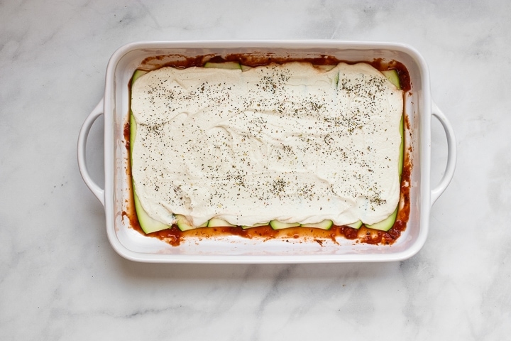 White baking dish with tomato sauce topped with zucchini slices and almond ricotta spread with Italian seasonings on gray marble. 