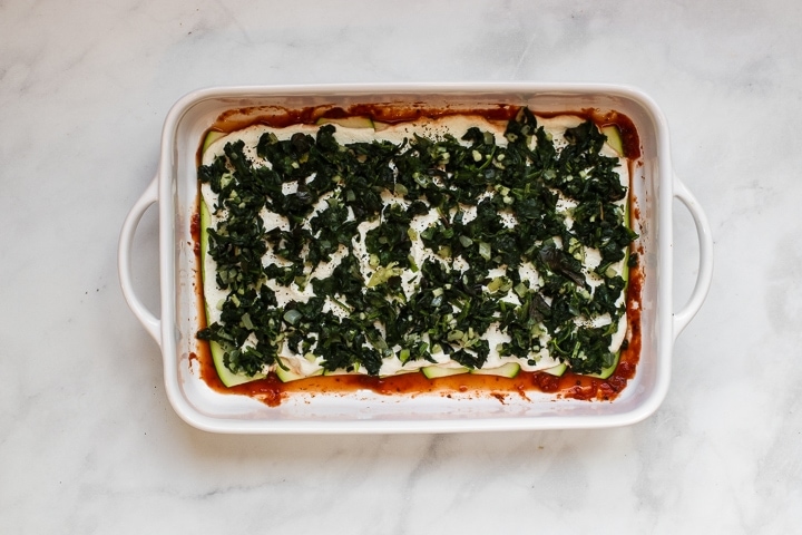 White baking dish with tomato sauce topped with zucchini slices and almond ricotta spread with Italian seasonings and spinach basil mixture on gray marble. 